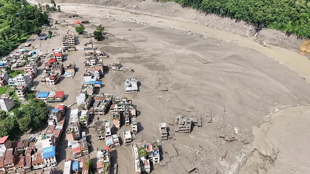 Aerial view of a Trishuli corridor town buried under grey flood sediment, buildings partially collapsed or submerged along the debris-choked riverbed