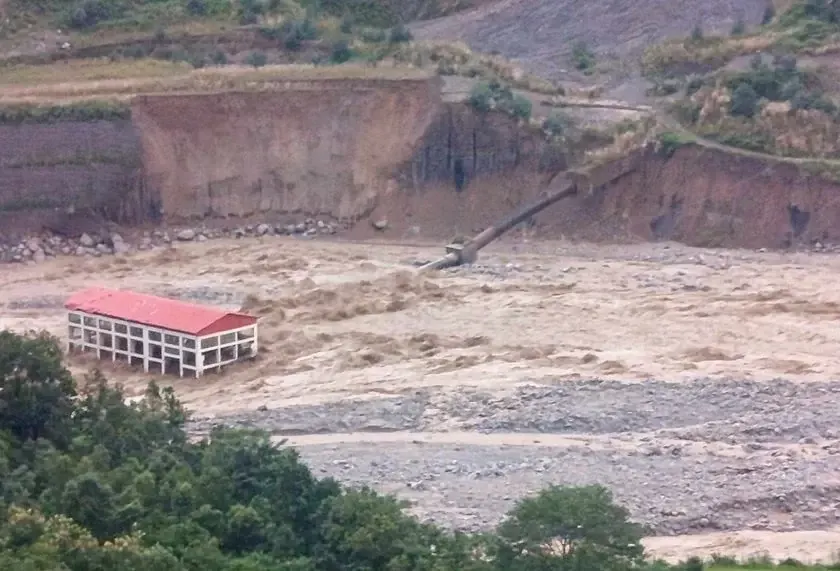A red roofed building nearly submerged in the flood, with a damaged penstock pipe angled into the debris filled river