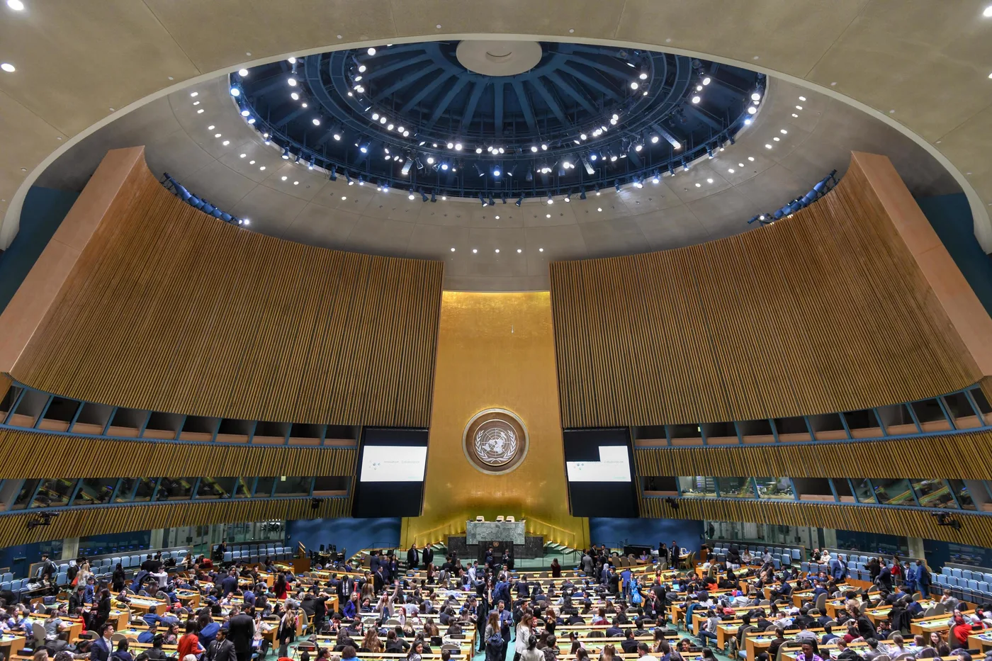 Interior of the United Nations General Assembly hall in New York, with delegates seated at desks below the podium and the UN emblem on the wall behind it